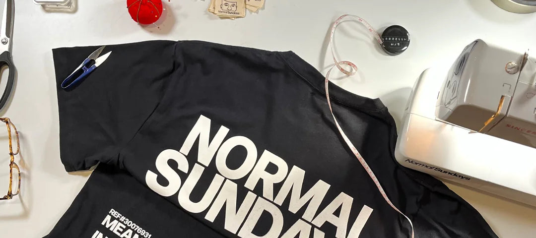 Black t-shirt with 'NORMAL SUNDAY' text on a white surface with sewing tools.Black t-shirt with 'NORMAL SUNDAY' text on a white surface with sewing tools.